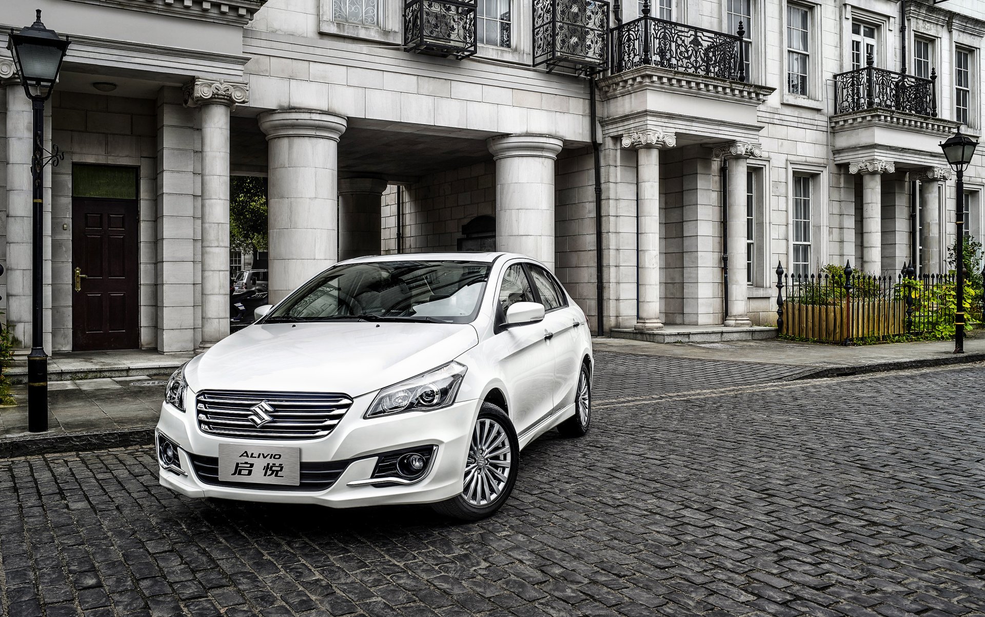 White Suzuki Ciaz compact car parked on cobblestone street in front of a neoclassical building — 2K Quad HD PC desktop wallpaper/background featuring a Suzuki vehicle.