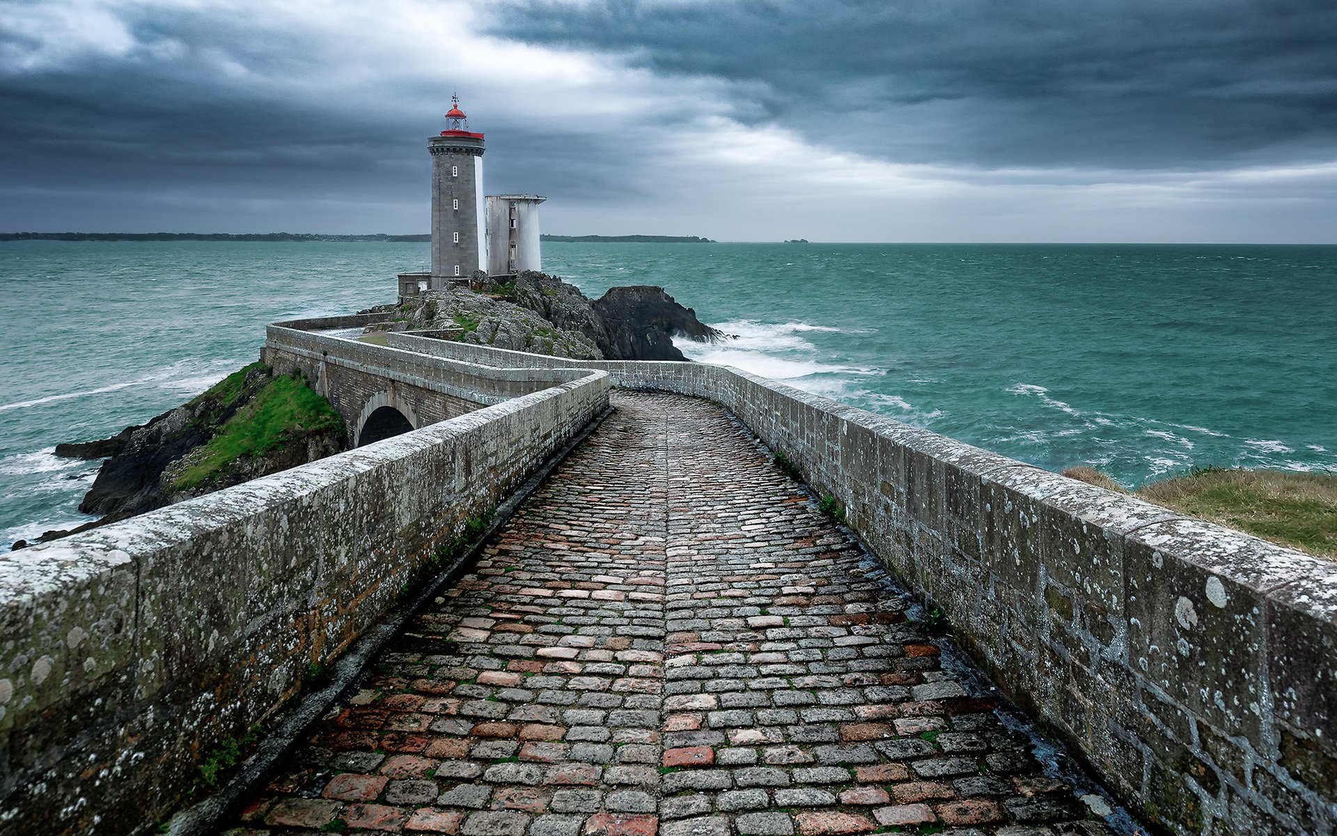 HD desktop wallpaper showing a stone walkway leading to a man-made lighthouse on a rocky coastline with the ocean and horizon under a cloudy sky.
