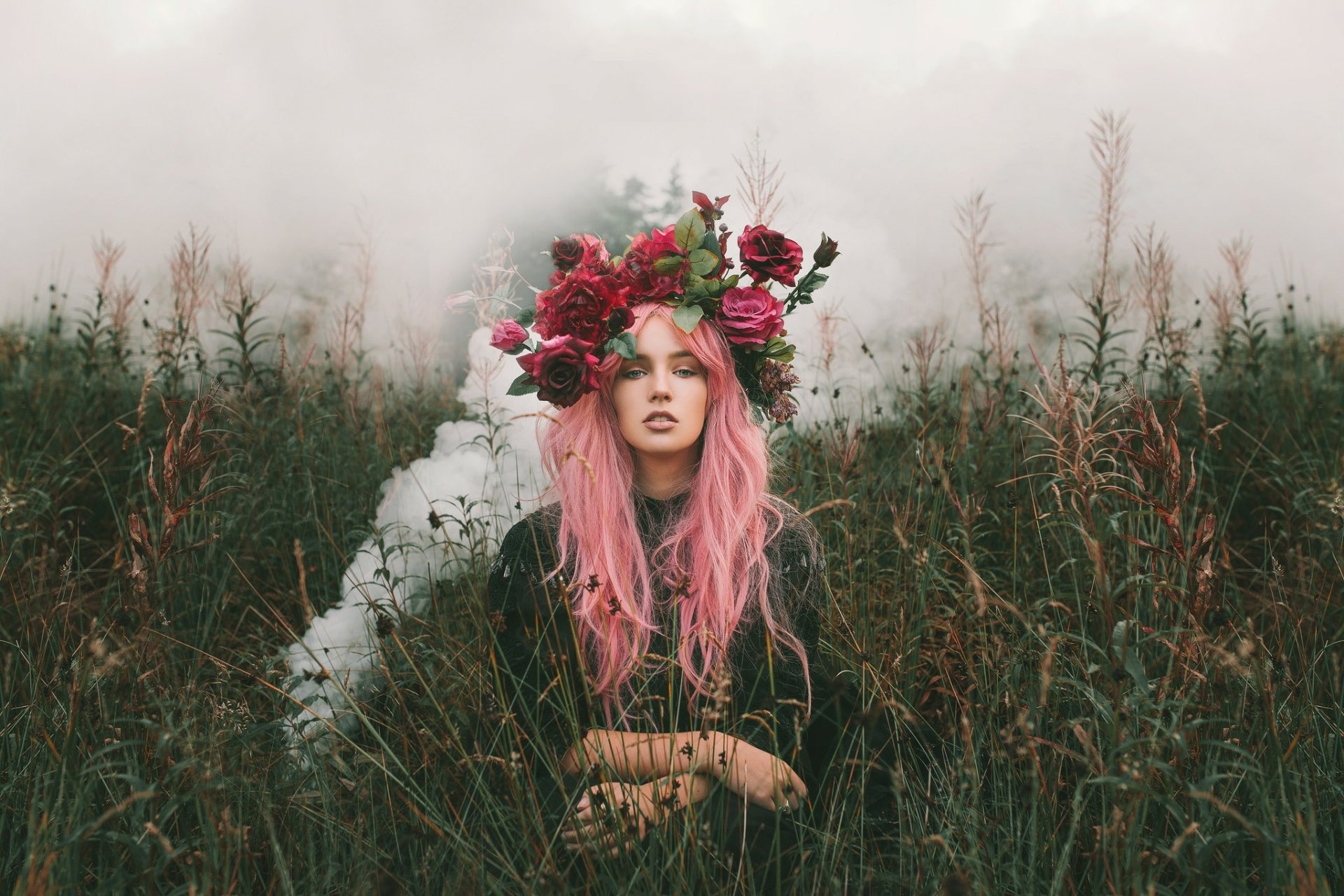 HD PC wallpaper of a woman with long pink hair and a flower wreath standing in a misty grassy field, flowers woven in her hair, creating a moody, ethereal background.