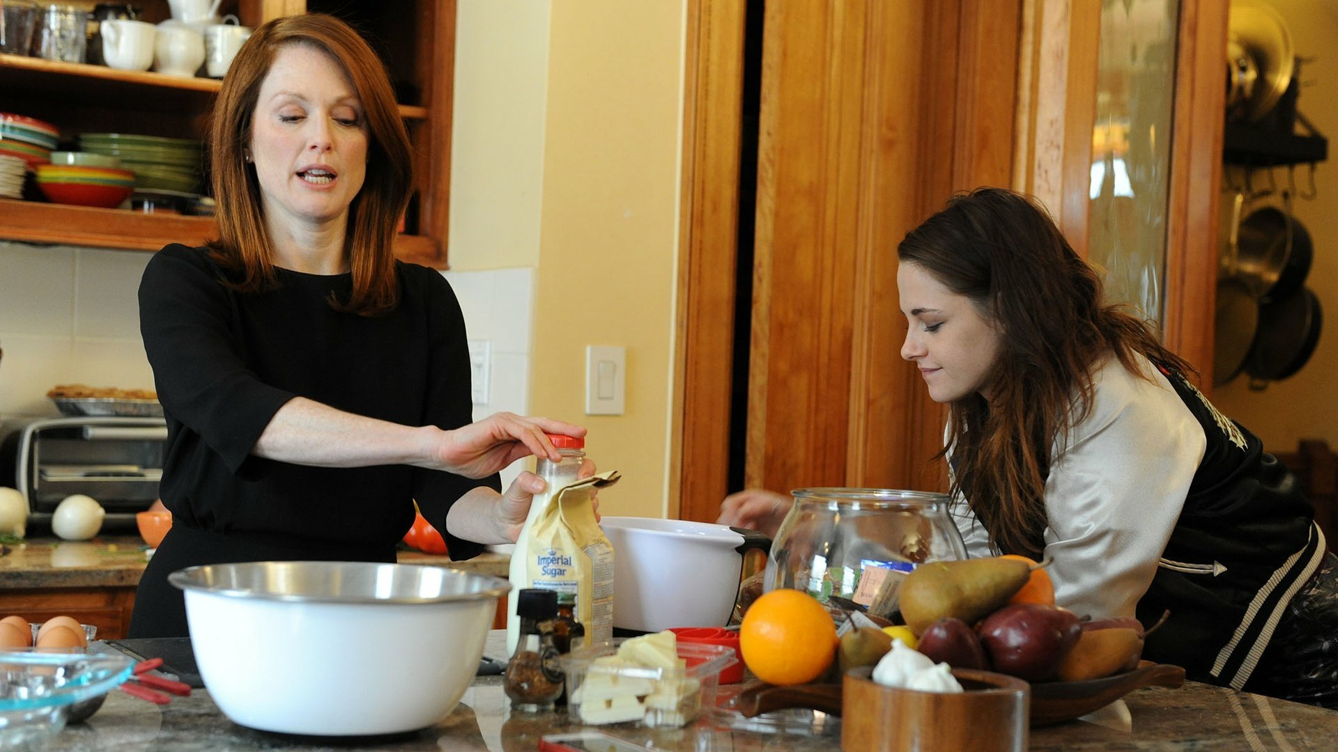 HD PC wallpaper featuring Kristen Stewart and Julianne Moore in a scene from the movie Still Alice, set in a kitchen with fruit and baking bowls on the counter.