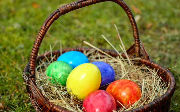 A rustic wicker basket filled with colorful Easter eggs resting on hay, set against a grassy background, captured in vibrant 4K Ultra HD for PC desktop wallpaper.