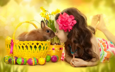 A cute little girl with a flower in her hair lies next to a yellow Easter basket with a bunny and colorful Easter eggs, creating a bright holiday HD desktop wallpaper.