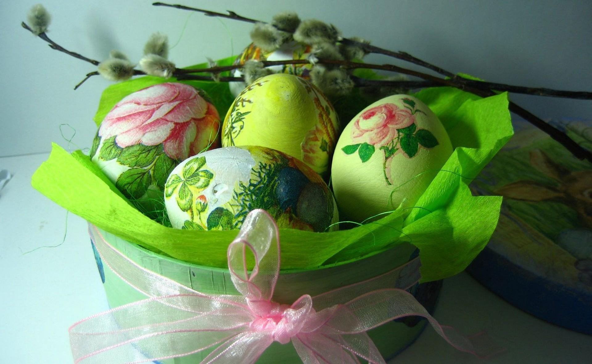 Colorful Easter eggs with floral designs nestled in a basket lined with green tissue paper, adorned with a pink ribbon and pussy willow branches in the background.