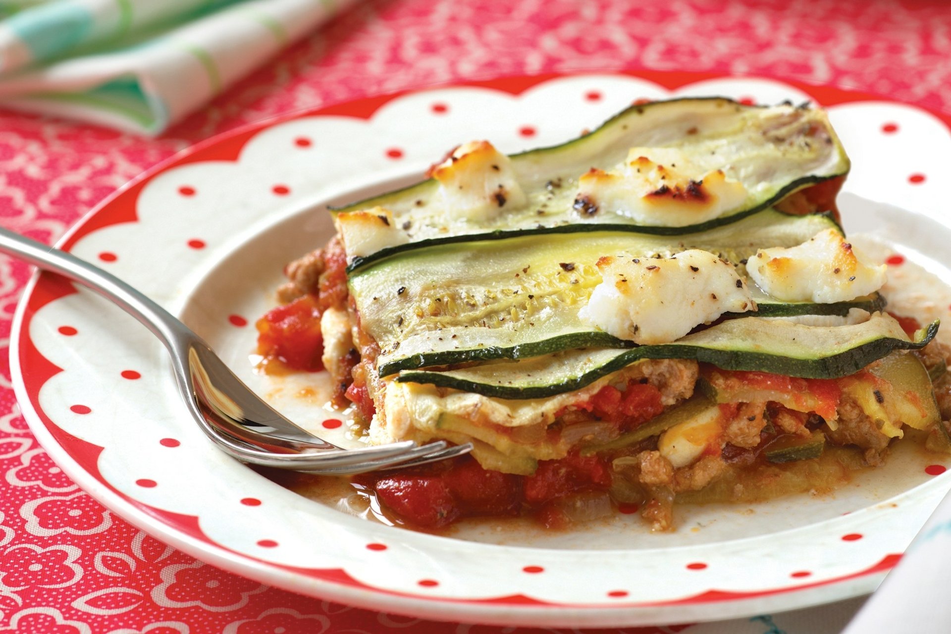 HD PC desktop wallpaper showing a plated lasagna meal: cheesy layered lasagna with tomato sauce and zucchini slices on a polka-dot plate with a fork.