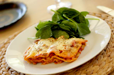 4K Ultra HD desktop wallpaper showing a plated slice of cheesy lasagna with tomato sauce and melted cheese, accompanied by fresh spinach on a white plate over a woven placemat.