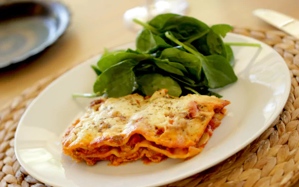 4K Ultra HD desktop wallpaper showing a plated slice of cheesy lasagna with tomato sauce and melted cheese, accompanied by fresh spinach on a white plate over a woven placemat.