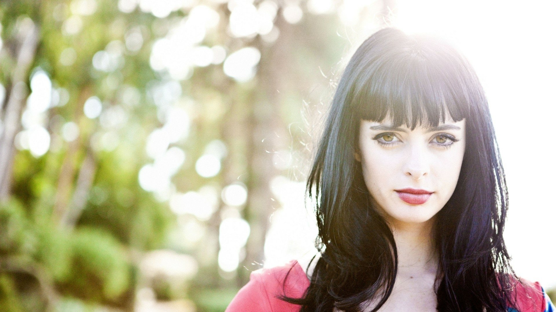 HD desktop wallpaper portrait of a woman with long dark hair and blunt bangs, pale skin and red lips, sunlit bokeh of green trees behind her.