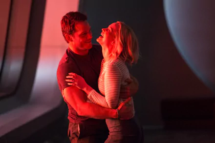HD PC desktop wallpaper showing two actors from the movie Passengers embracing and laughing in a red-lit spacecraft cabin beside a curved window.