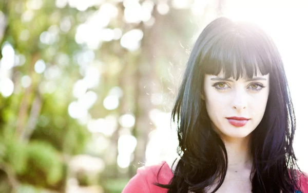 HD desktop wallpaper portrait of a woman with long dark hair and blunt bangs, pale skin and red lips, sunlit bokeh of green trees behind her.