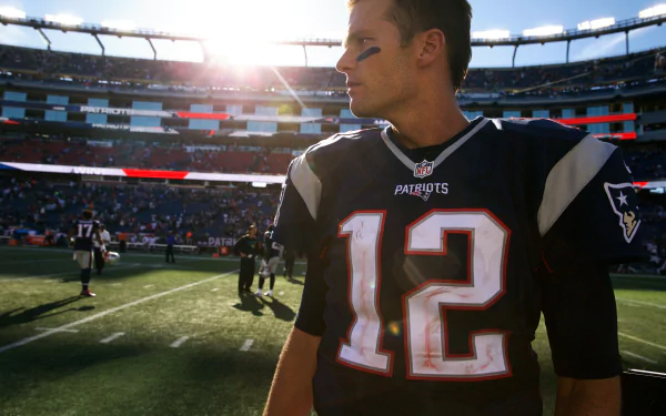 2K Quad HD PC wallpaper showing a New England Patriots quarterback (#12) on the field at sunset with the stadium crowd behind him.