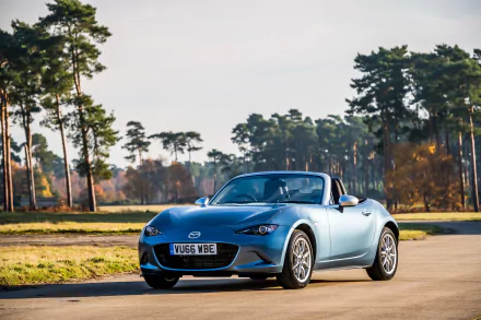 A sleek Mazda MX-5 convertible in metallic blue parked on a sunlit road surrounded by pine trees, captured in high-resolution 4K Ultra HD for desktop background use.