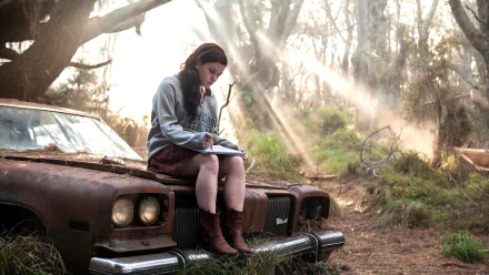 A scene from Evil Dead (2013) featuring a girl sitting on an old car, immersed in writing, with rays of sunlight piercing through the forest behind her. HD desktop wallpaper and background.