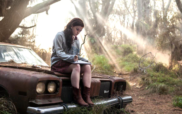 A scene from Evil Dead (2013) featuring a girl sitting on an old car, immersed in writing, with rays of sunlight piercing through the forest behind her. HD desktop wallpaper and background.