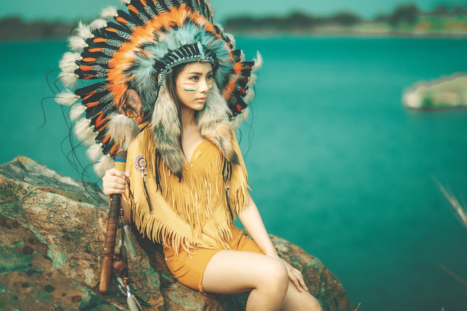 HD desktop wallpaper of an Asian woman model wearing a Native American feather headdress, sitting on a rock by a turquoise body of water with a shallow depth of field.