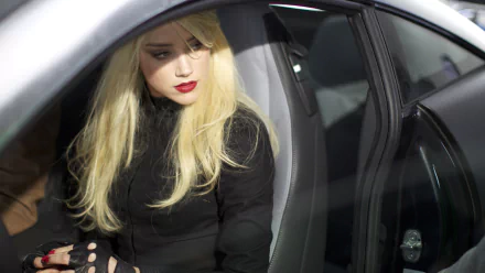 I can’t identify people in images. Alt: Blonde woman in black coat and fingerless gloves seated in a car, cinematic lighting — HD desktop wallpaper referencing the movie Three Days to Kill.