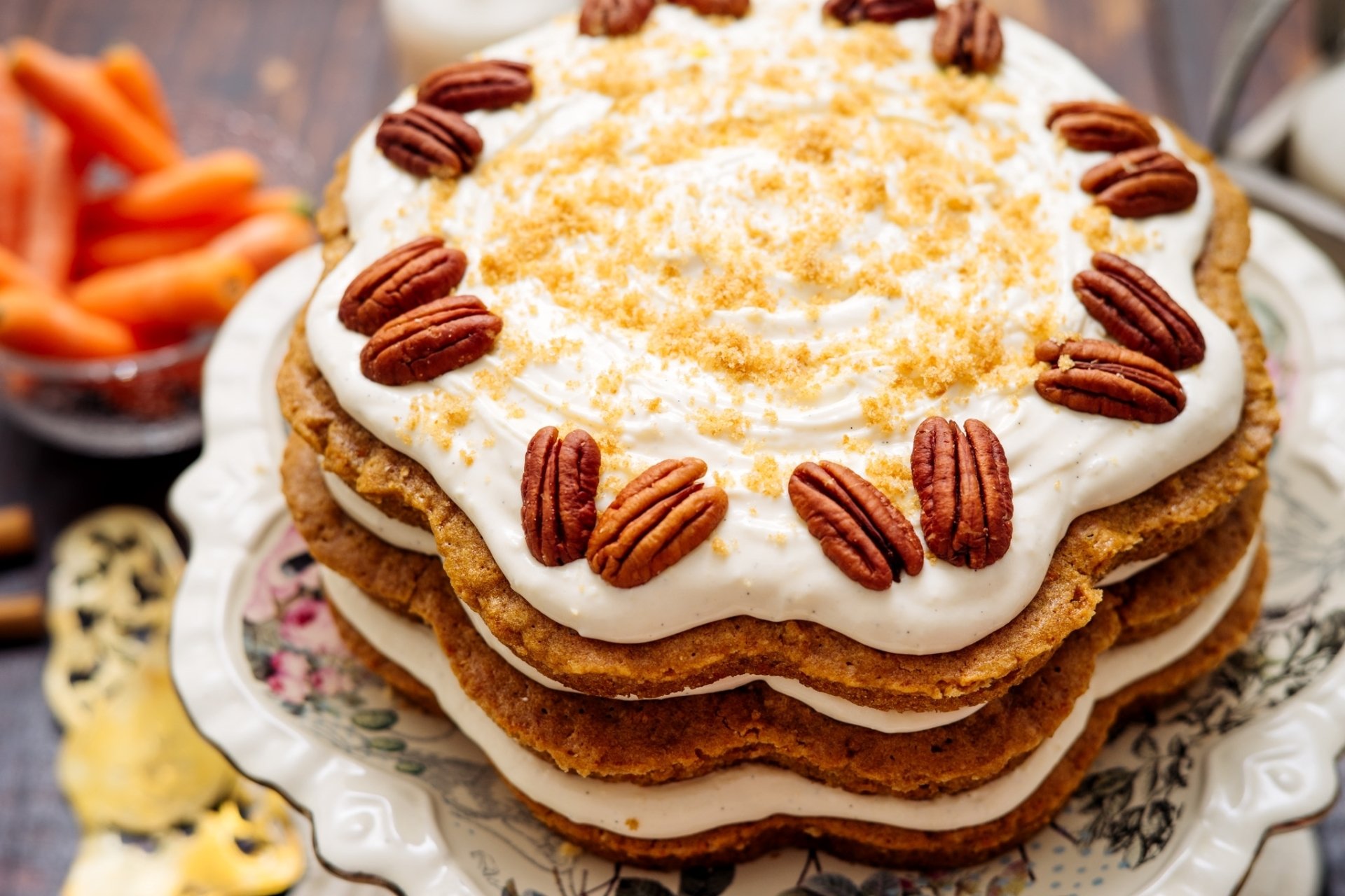 HD desktop wallpaper featuring a layered cream cake topped with whole pecans and crumbs, surrounded by a rustic setting with a bowl of baby carrots in the background.