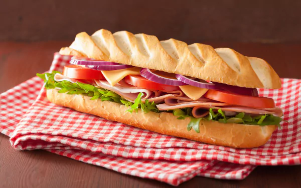 4K Ultra HD desktop wallpaper featuring a fresh sandwich with layers of lettuce, tomato, onion, cheese, and deli meat on a crusty baguette over a red checkered cloth.