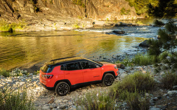 HD desktop wallpaper of a red Jeep Compass SUV parked by a rocky riverbank surrounded by trees and sunlight.
