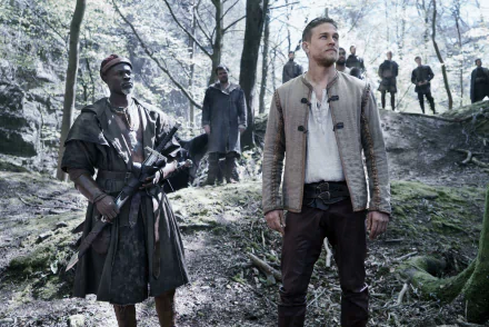 HD wallpaper featuring Djimon Hounsou and Charlie Hunnam as characters in King Arthur: Legend of the Sword, set in a forest with additional figures in the background.