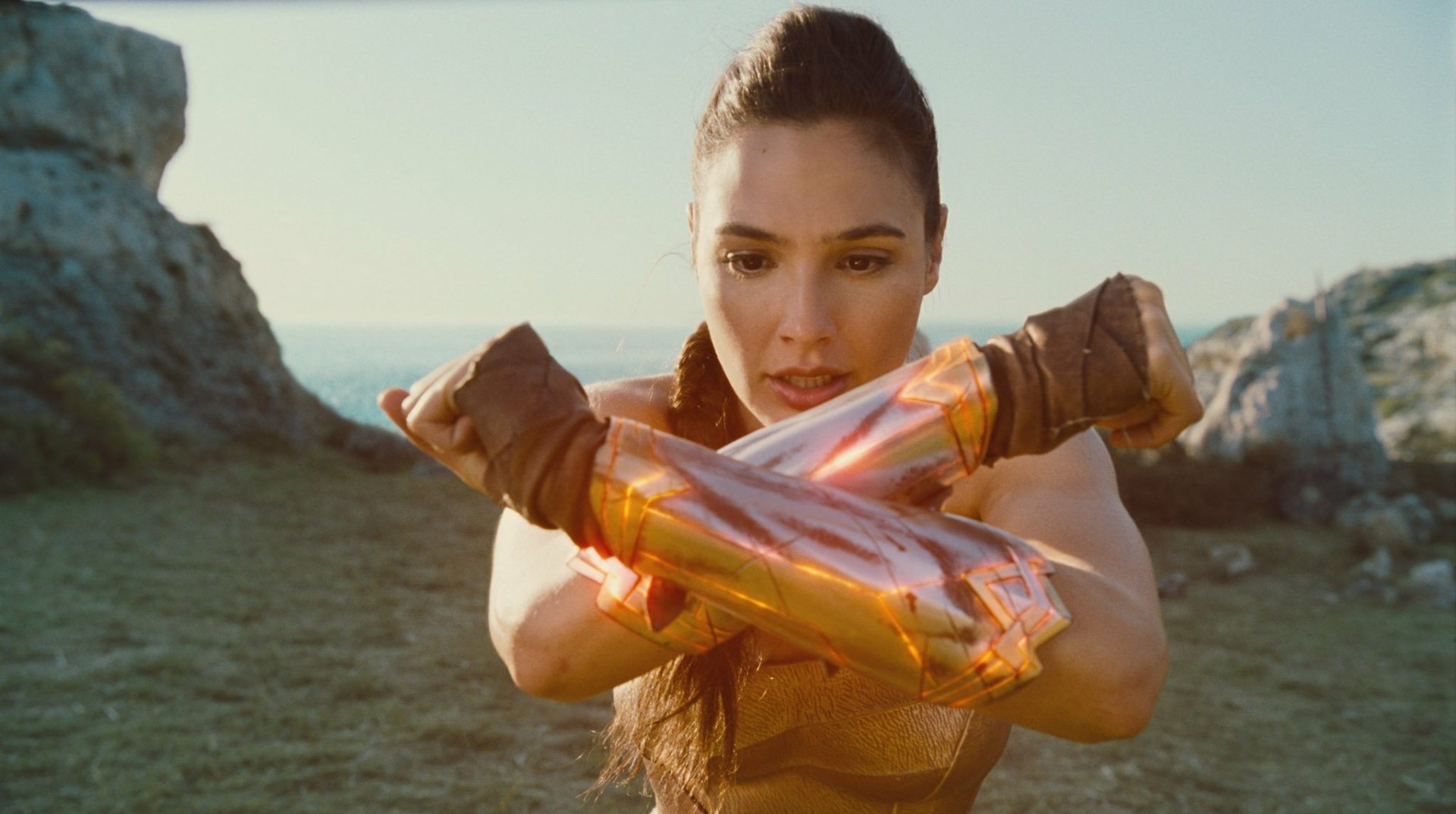 HD desktop wallpaper of Gal Gadot as Wonder Woman, crossing her glowing gauntlets in a powerful stance, set against a rugged outdoor backdrop.