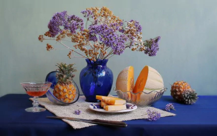 HD still life photography of melon slices, a whole melon, pineapple, and dried flowers arranged in a blue vase on a blue tablecloth, designed as a PC desktop wallpaper.