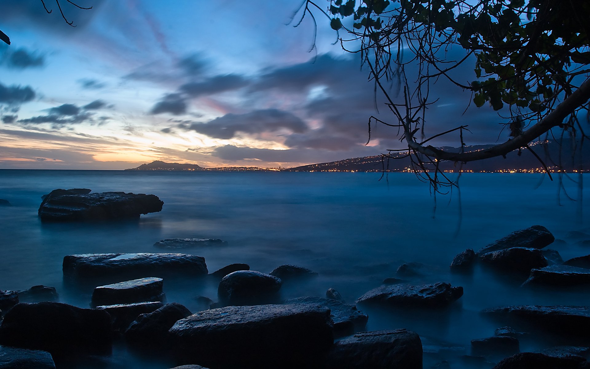A serene lakeside view at dusk, showcasing rocks in the foreground, a cloudy sky, and the city skyline illuminated in the background, creating a harmonious blend of nature and urban life.