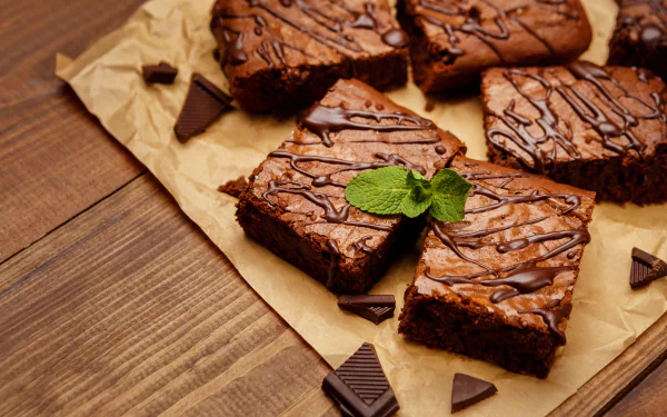 Close-up still life of chocolate brownies drizzled with chocolate, garnished with mint leaves, arranged on parchment paper with chocolate pieces on a wooden surface in 4K Ultra HD.