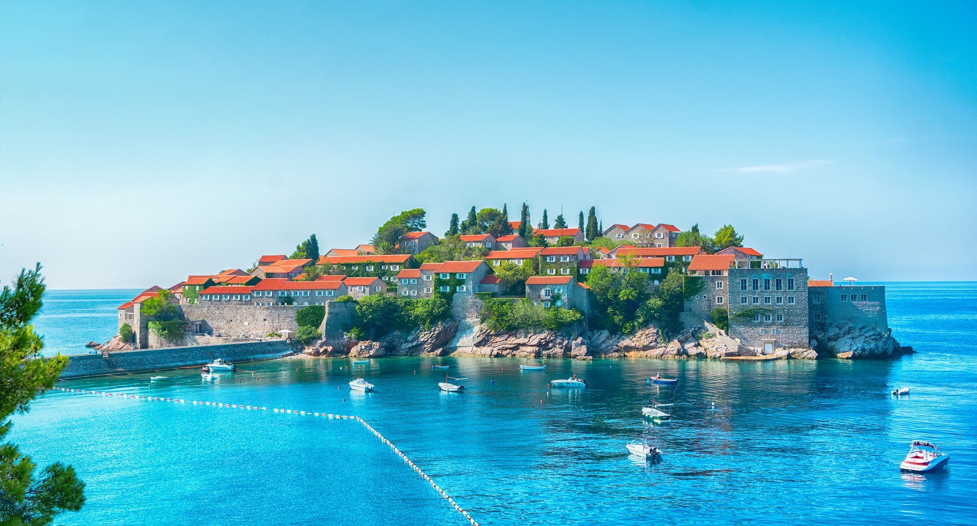 A man-made island village in Montenegro with stone houses featuring red roofs, surrounded by boats and clear blue water under a bright sky.