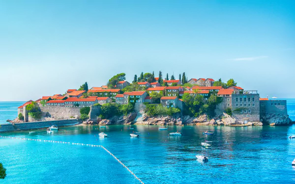 A man-made island village in Montenegro with stone houses featuring red roofs, surrounded by boats and clear blue water under a bright sky.