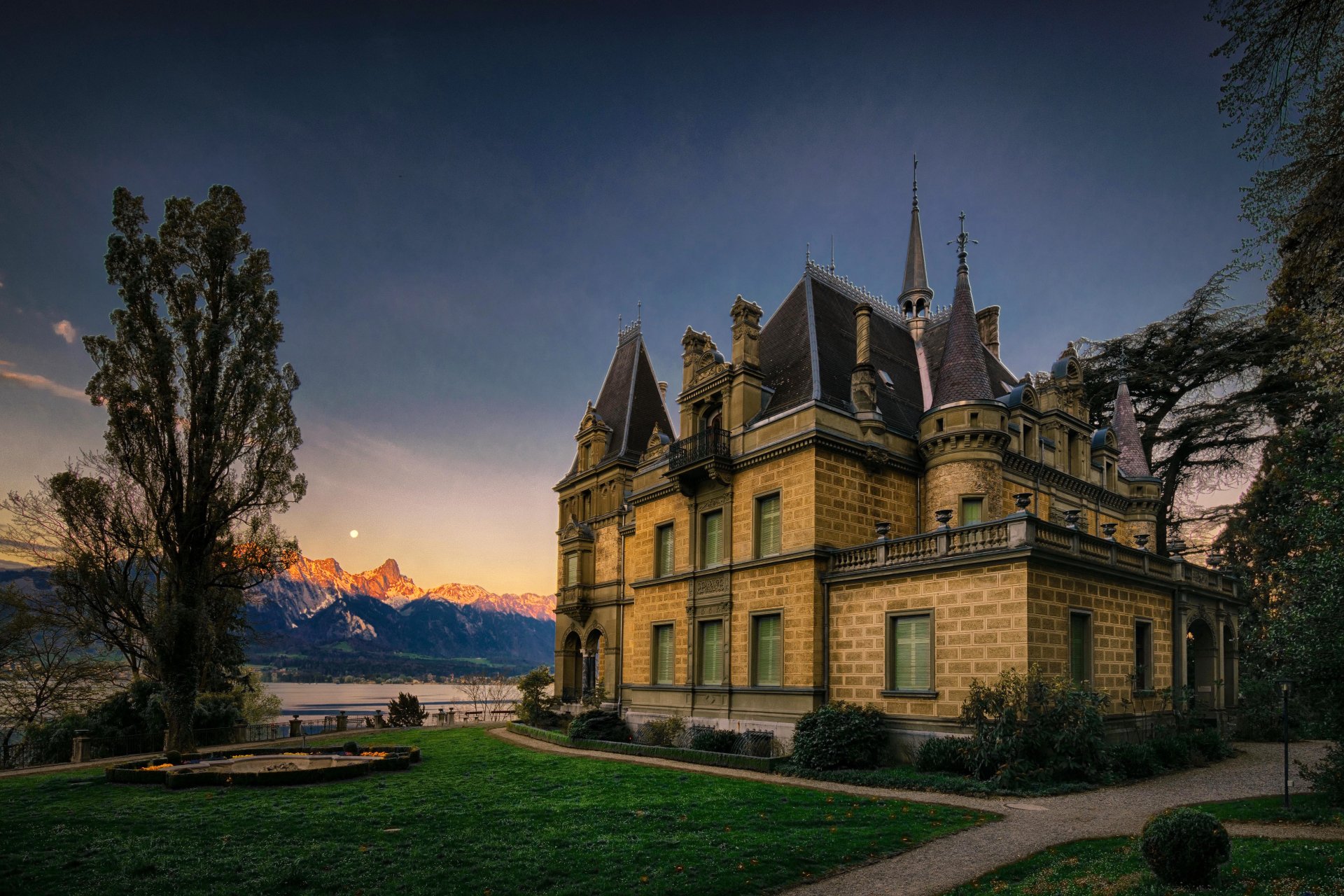 Hunegg Castle in Switzerland stands against a twilight sky, showcasing intricate architecture with mountain peaks and a lake in the background.