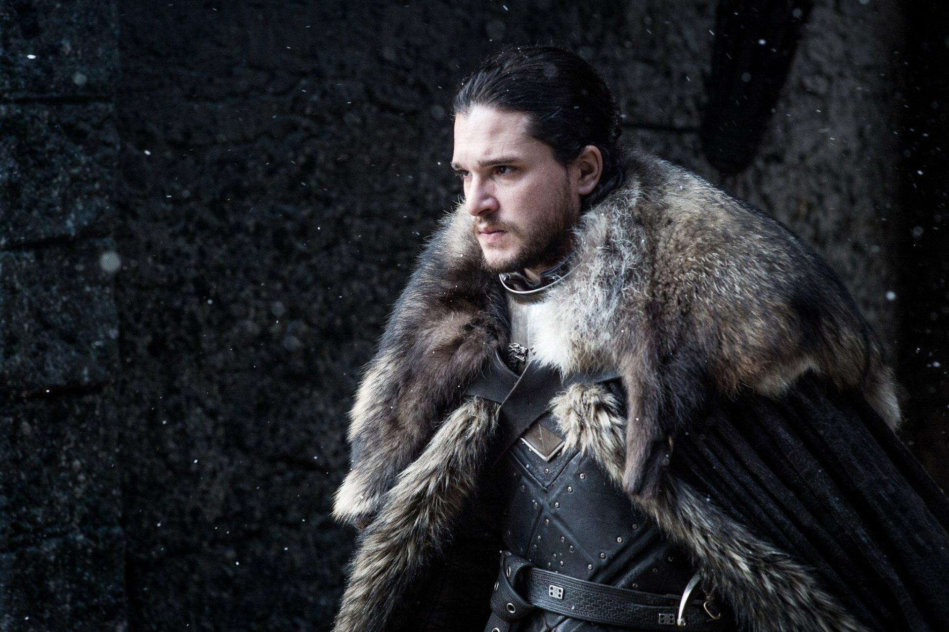 Jon Snow, portrayed by Kit Harington in Game of Thrones, stands solemnly in dark medieval armor with a heavy fur cloak against a stone wall background.