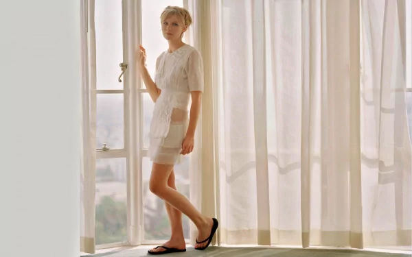 A high-definition wallpaper featuring Kirsten Dunst standing gracefully by a window, dressed in a stylish outfit with soft light filtering through sheer curtains.