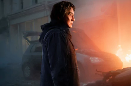 A HD desktop wallpaper featuring Jackie Chan in a dramatic scene from the movie The Foreigner, with a car and a fiery background.