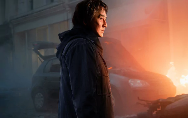 A HD desktop wallpaper featuring Jackie Chan in a dramatic scene from the movie The Foreigner, with a car and a fiery background.