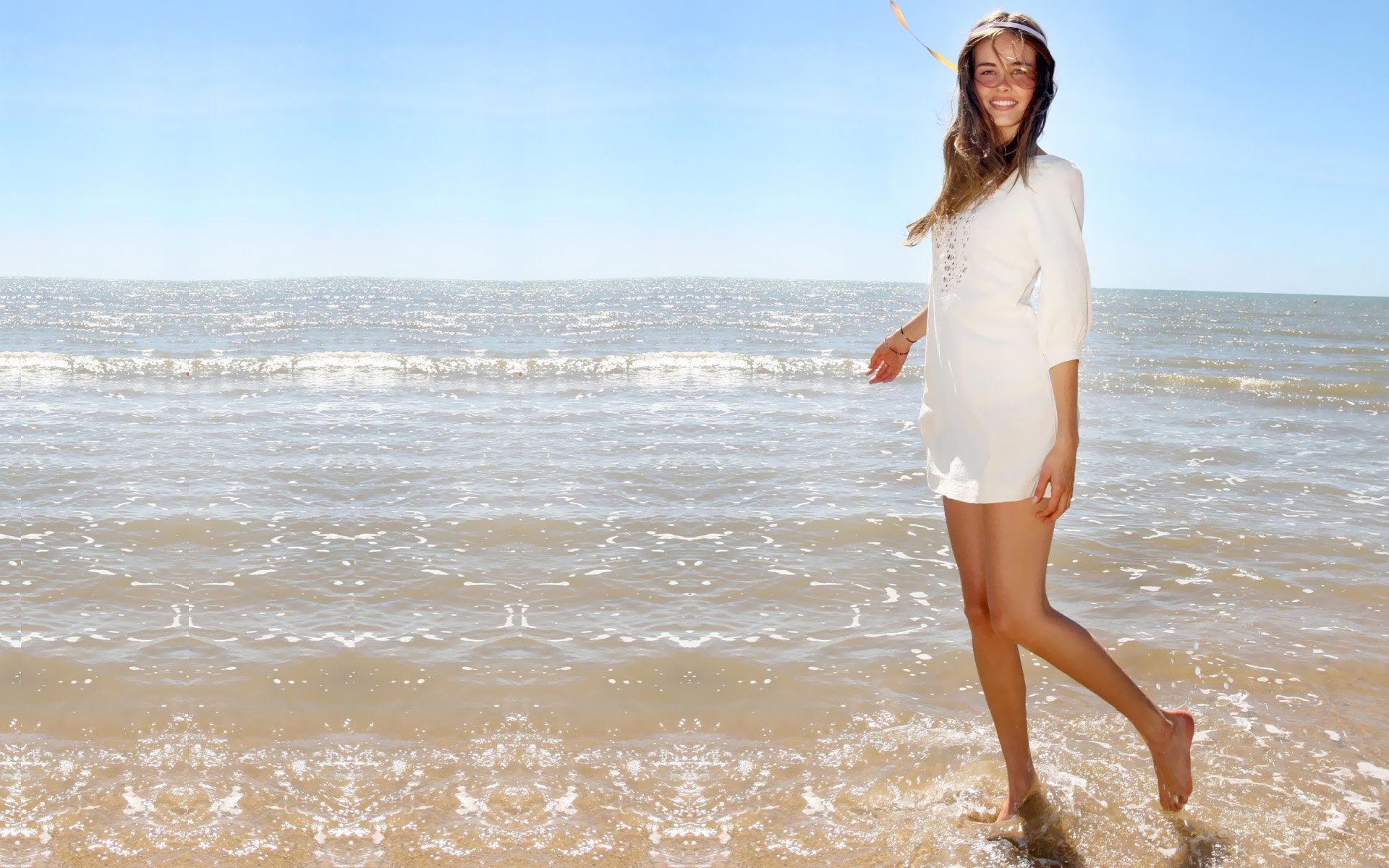 Isabel Lucas joyfully walks along the beach, her feet in the water, with the sun shining and waves gently lapping at the shore, creating a serene and vibrant HD desktop wallpaper.