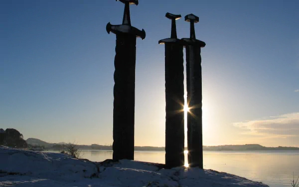 HD PC desktop wallpaper featuring a man-made statue of three large swords standing upright against a serene lakeside sunset background.
