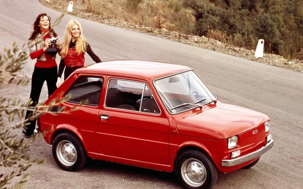 Two women stand beside a red Fiat 126 on a winding road, surrounded by greenery. This HD desktop wallpaper showcases vintage automotive charm in a scenic outdoor setting.