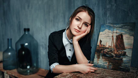 HD wallpaper of a brunette model with brown eyes, Vasilisa Sarovskaya, resting her head on her hand near a painting of a boat. Background features a rustic, artistic indoor setting.