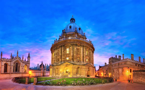 HD PC desktop wallpaper of Radcliffe Camera at Oxford University, Britain, lit at dusk with parked bicycles in front, historic man-made rotunda and surrounding college buildings.