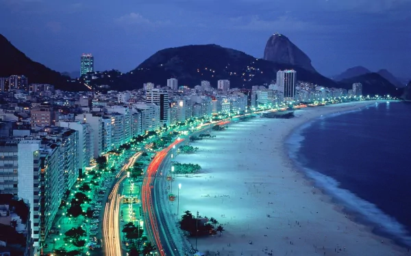 HD wallpaper and background image of Rio de Janeiro at night, featuring a beautifully illuminated beach with city lights and buildings stretching along the coastline under a twilight sky.
