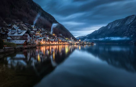Nighttime view of the charming town of Hallstatt, Austria, with illuminated houses reflecting on the calm lake, surrounded by majestic mountains under a moody sky.
