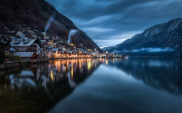 Nighttime view of the charming town of Hallstatt, Austria, with illuminated houses reflecting on the calm lake, surrounded by majestic mountains under a moody sky.