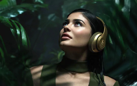 Close-up of American model Kylie Jenner with brown eyes and brunette hair wearing gold headphones, set against a lush green background in 4K Ultra HD quality.
