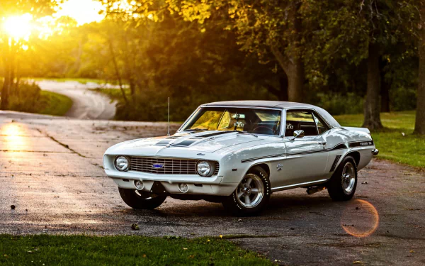 HD desktop wallpaper featuring a classic Chevrolet Camaro parked on a serene road at sunset, surrounded by lush greenery.