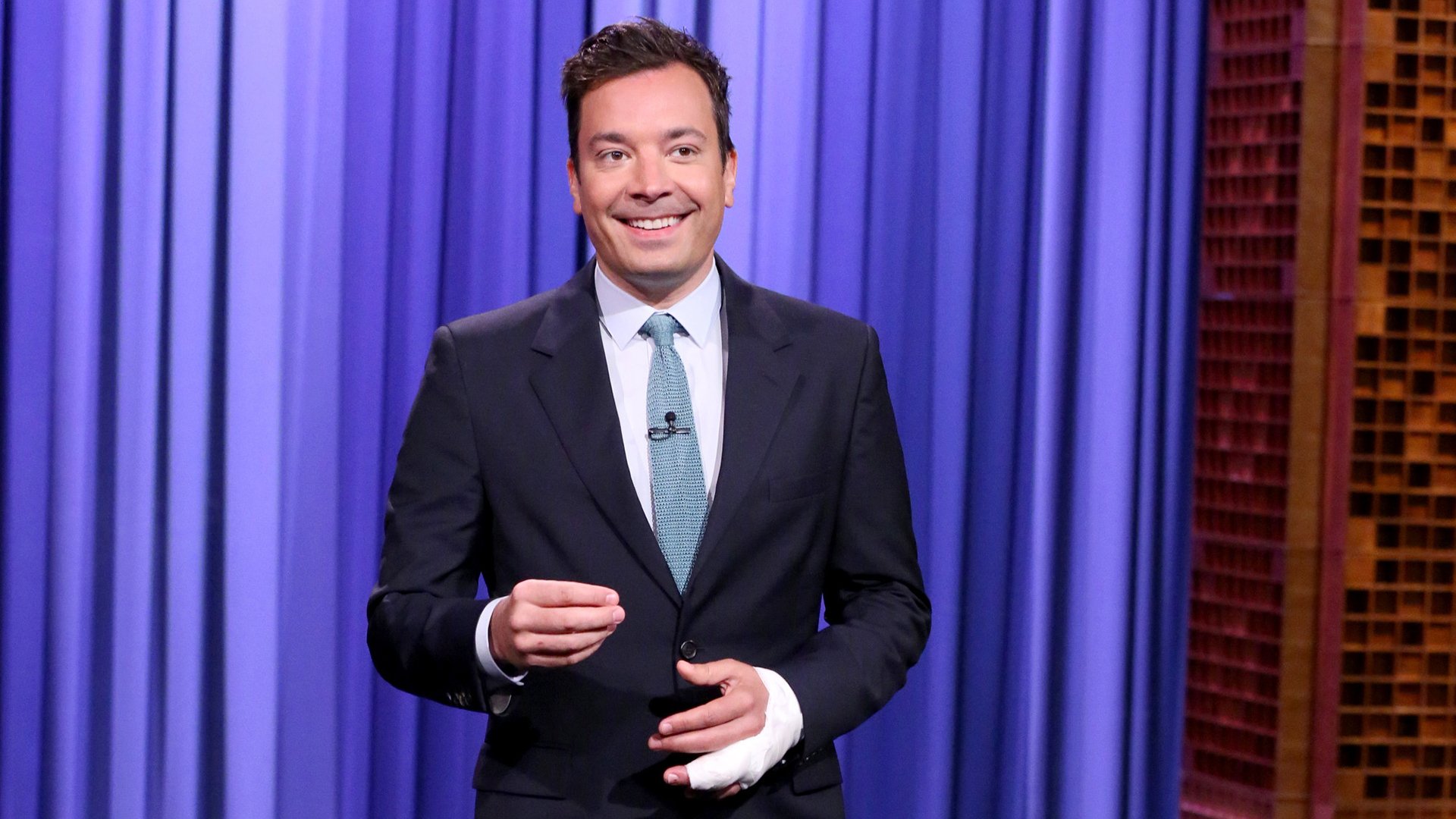 HD desktop wallpaper of the host on the set of The Tonight Show Starring Jimmy Fallon, featuring a smiling man in a suit with a blue curtain backdrop.