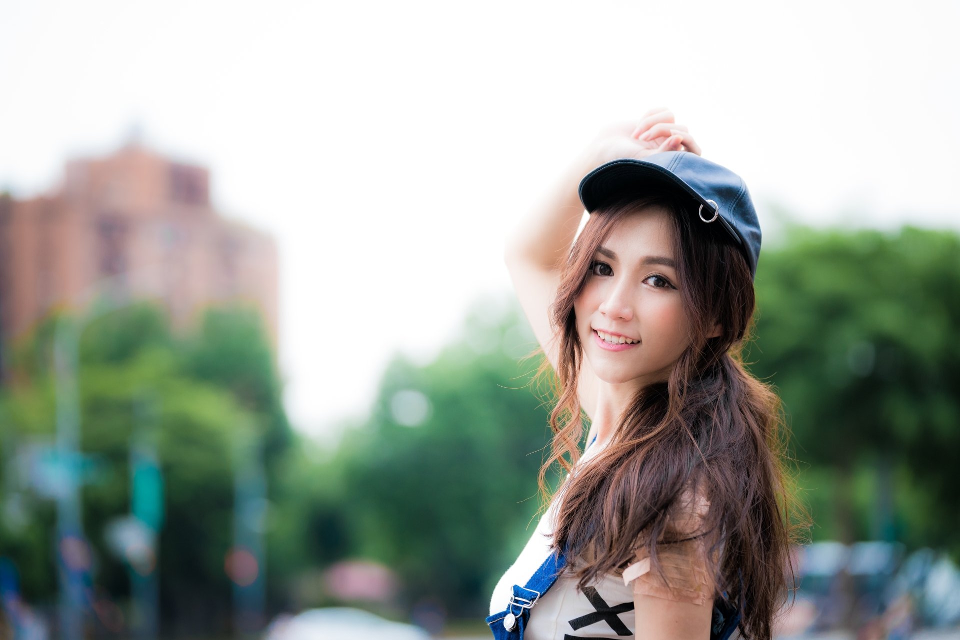 HD desktop wallpaper featuring a smiling brunette Asian woman in a cap. The image has a depth of field effect, with a soft-focus background of urban and green elements.