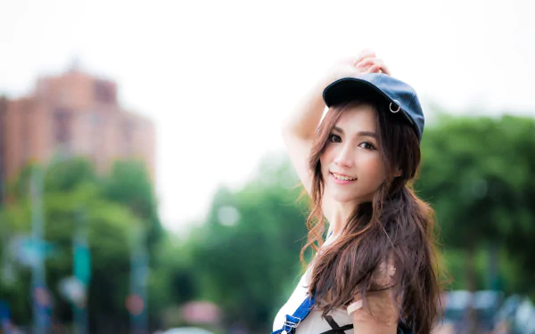 HD desktop wallpaper featuring a smiling brunette Asian woman in a cap. The image has a depth of field effect, with a soft-focus background of urban and green elements.