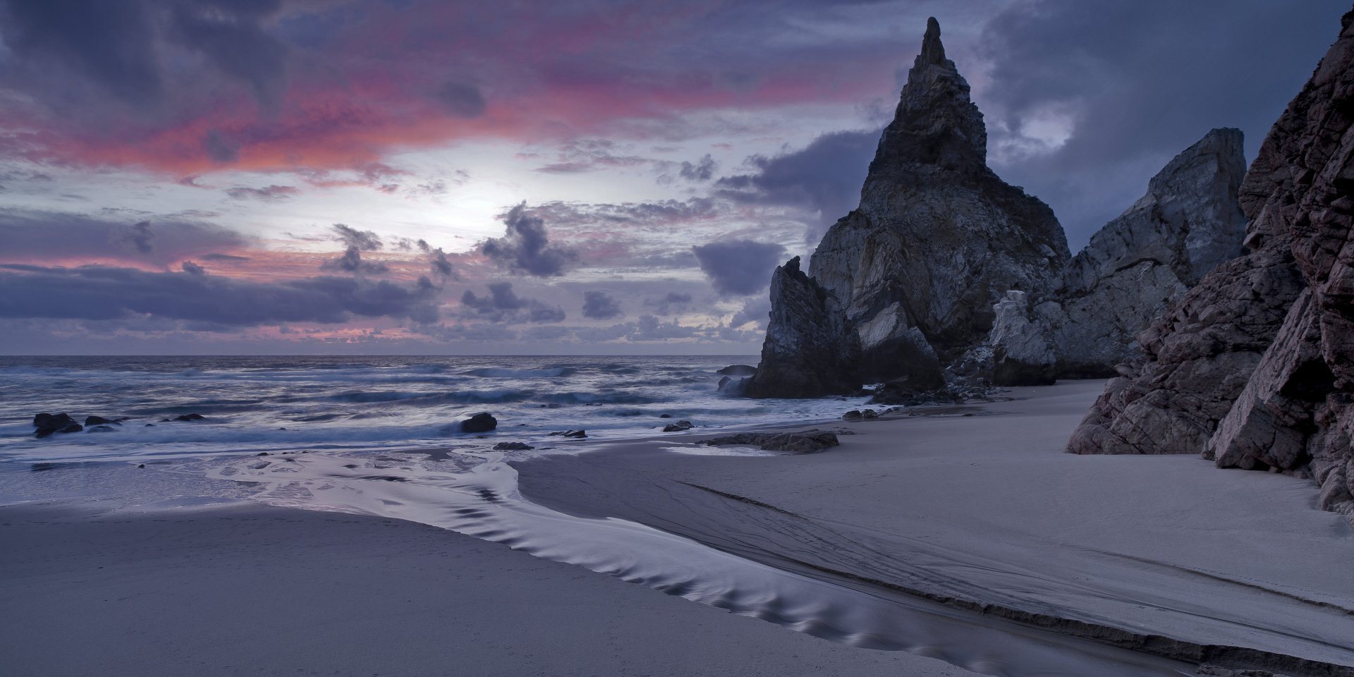 A stunning beach at sunset with dramatic clouds in a colorful sky, gentle waves, and sea cliffs. This HD desktop wallpaper captures the serene beauty of nature with the horizon in the distance.