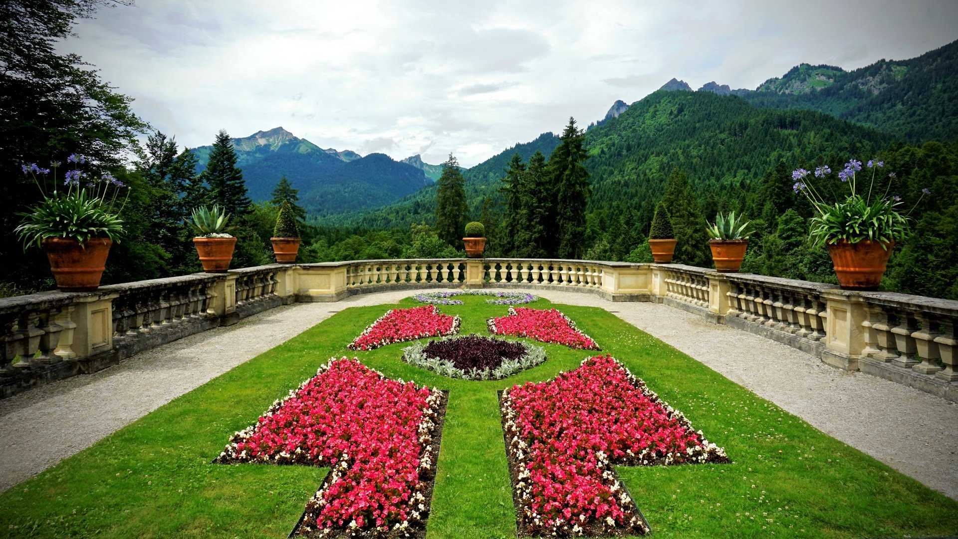 A vibrant garden with red flower beds arranged symmetrically in the courtyard of Linderhof Palace, with mountainous scenery in the background and a decorative fence lining the area.