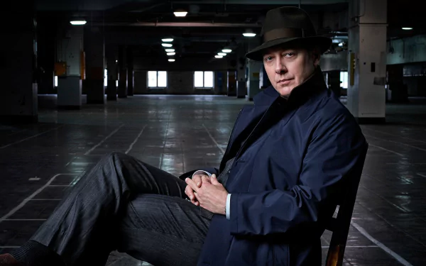 Raymond Reddington from The Blacklist sits in a dimly lit, empty industrial space, wearing a dark coat and fedora in this HD PC desktop wallpaper.